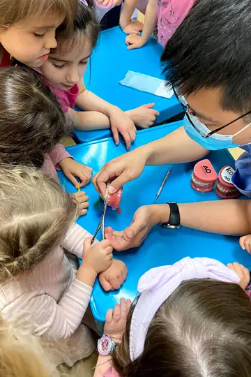 Several young children sit and stand around a blue table, watching an adult in a mask and glasses demonstrate brushing a red model tooth with a toothbrush.