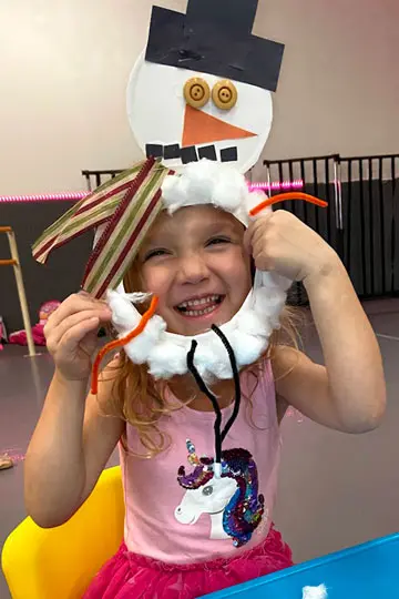 A smiling girl holds a paper snowman craft with a black hat, orange nose, and cotton-ball trim in front of her face.
