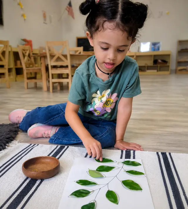 A child sits on a striped rug and places a green leaf shape onto a paper with a drawn branch while a small wooden bowl rests nearby.