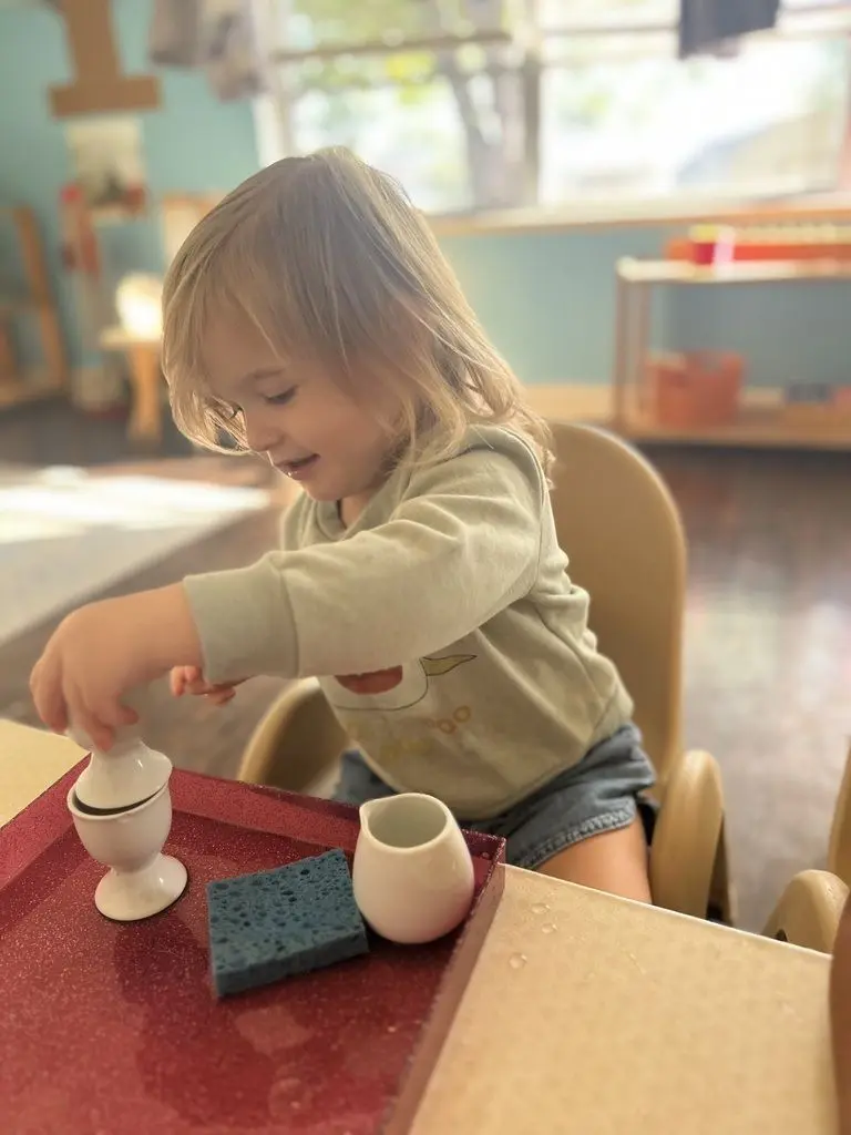 A young child with blonde hair sits at a table and holds a white egg cup over a red tray containing a sponge and a small pitcher.