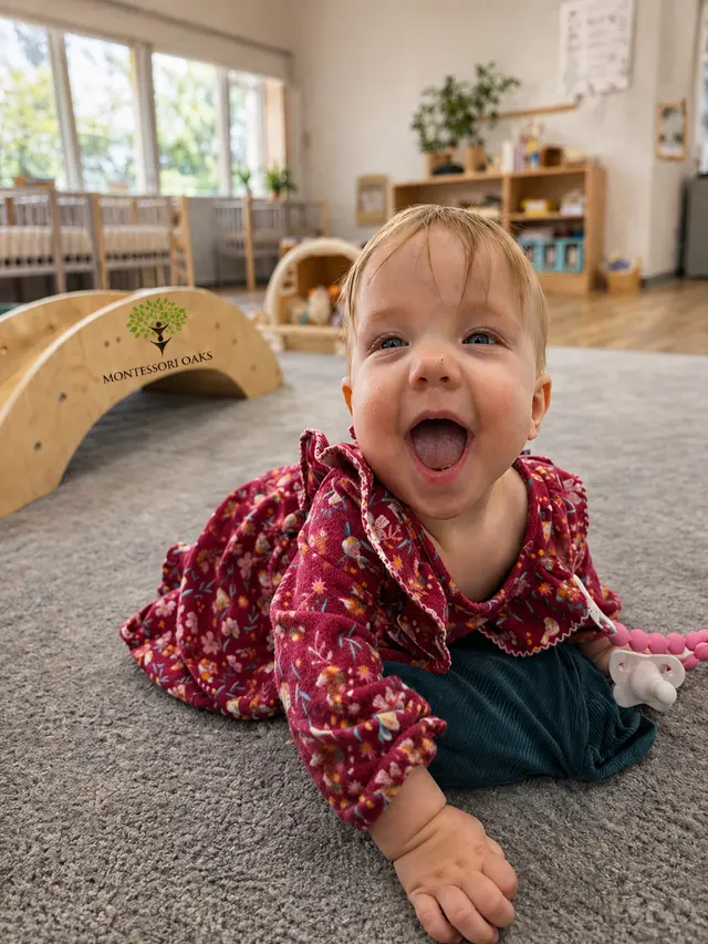 A baby in a floral dress lies on a gray rug, smiling with an open mouth in a room with wooden furniture and a climbing arch.