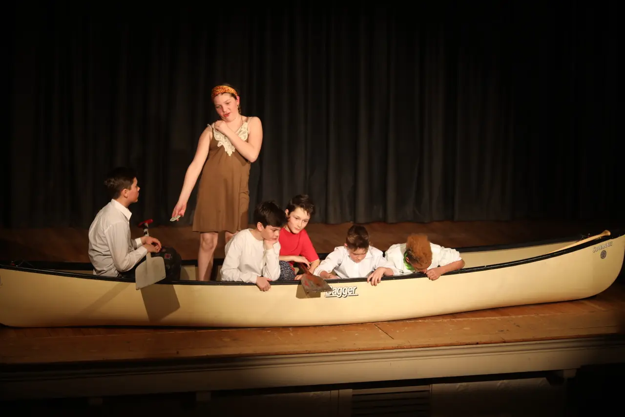 Six children perform on a stage, with one girl standing in a brown dress and five others seated or leaning inside a large beige canoe.