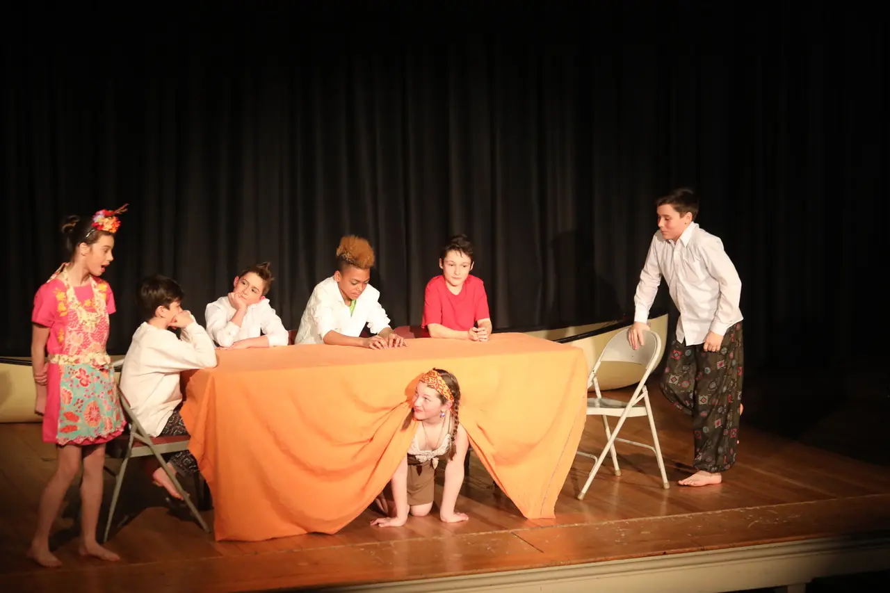 Seven children in costumes perform a scene around a table with an orange tablecloth on a stage, with one child crouching underneath the table.
