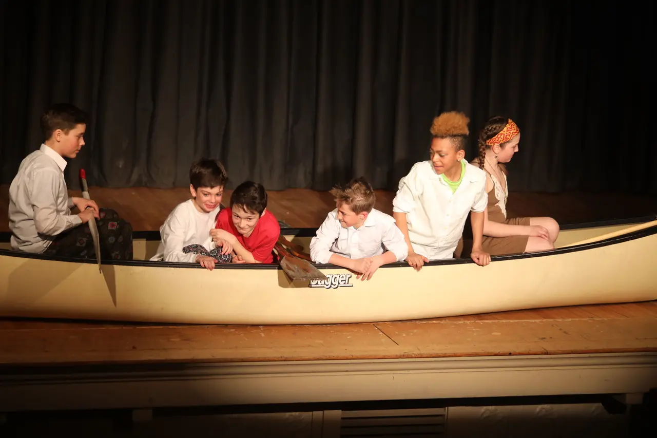 Six children sit inside a large beige canoe prop on a stage, with one boy holding a paddle and others leaning on the sides.