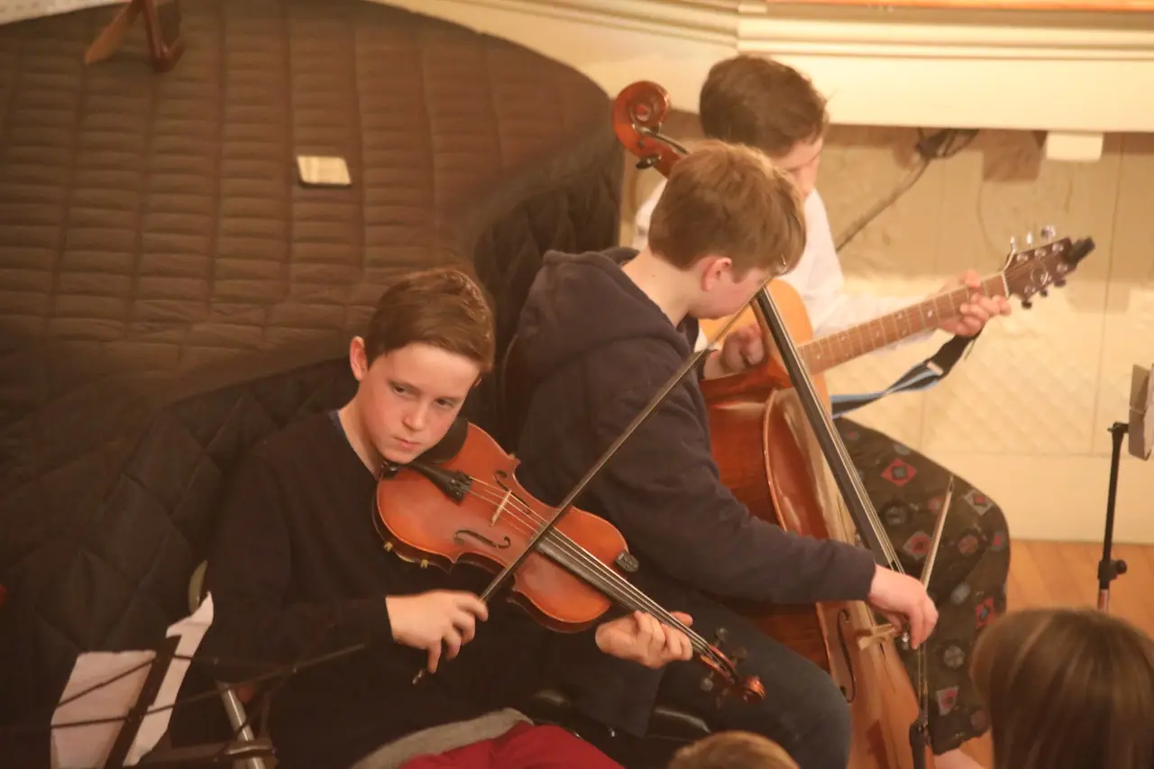 Three boys play string instruments including a violin, cello and guitar while seated indoors, with the top of another person's head visible in the foreground.