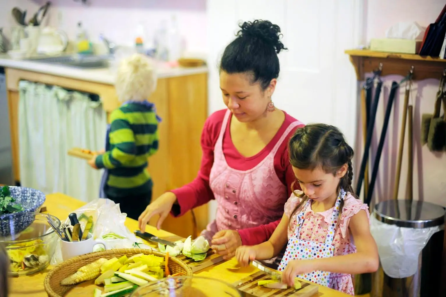 A woman in a pink apron and a young girl in a floral apron cut vegetables at a wooden counter while a child in a green striped shirt stands near cabinets in the background.