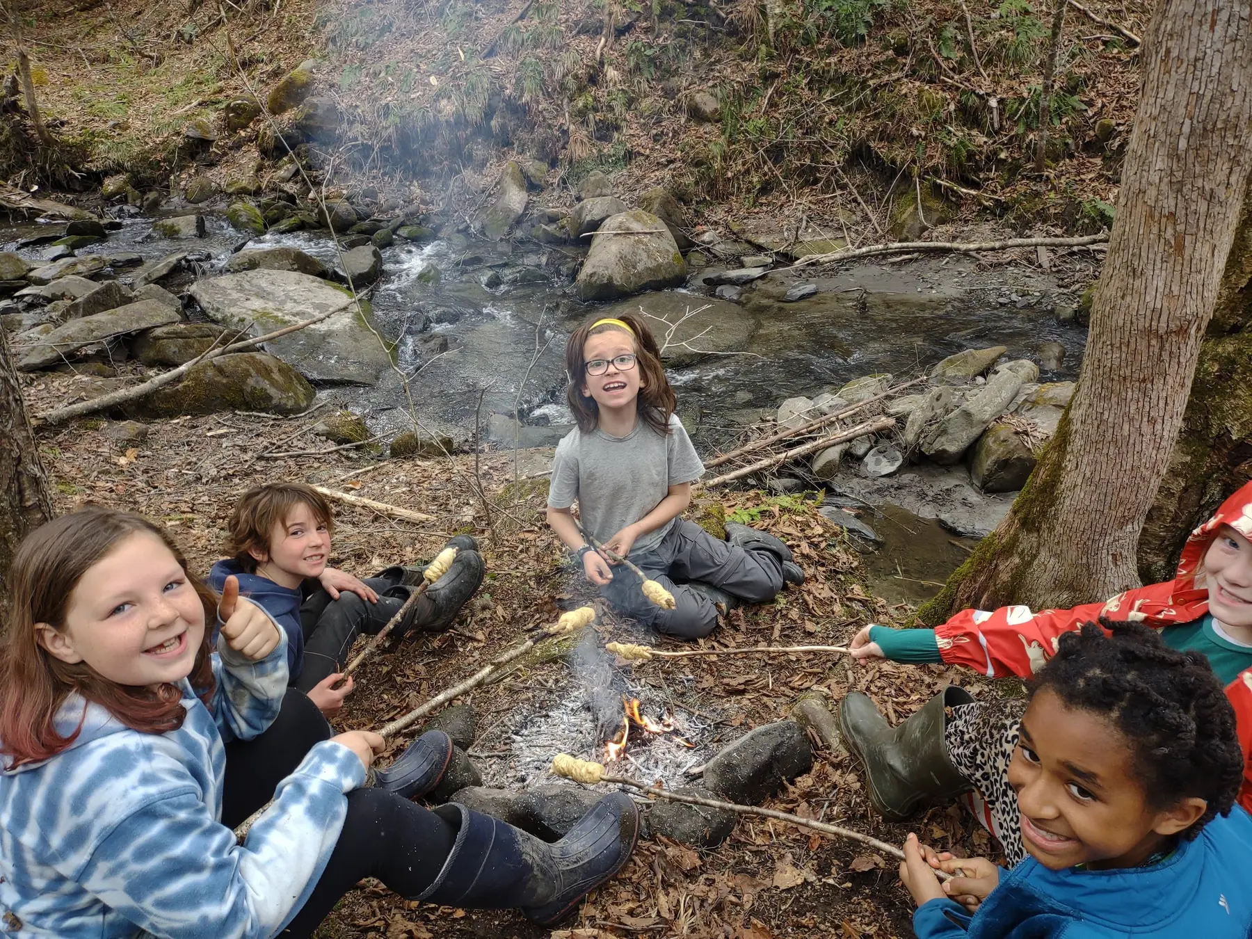 Five children sit and lie near a small campfire beside a rocky stream, holding sticks with food over the flames.