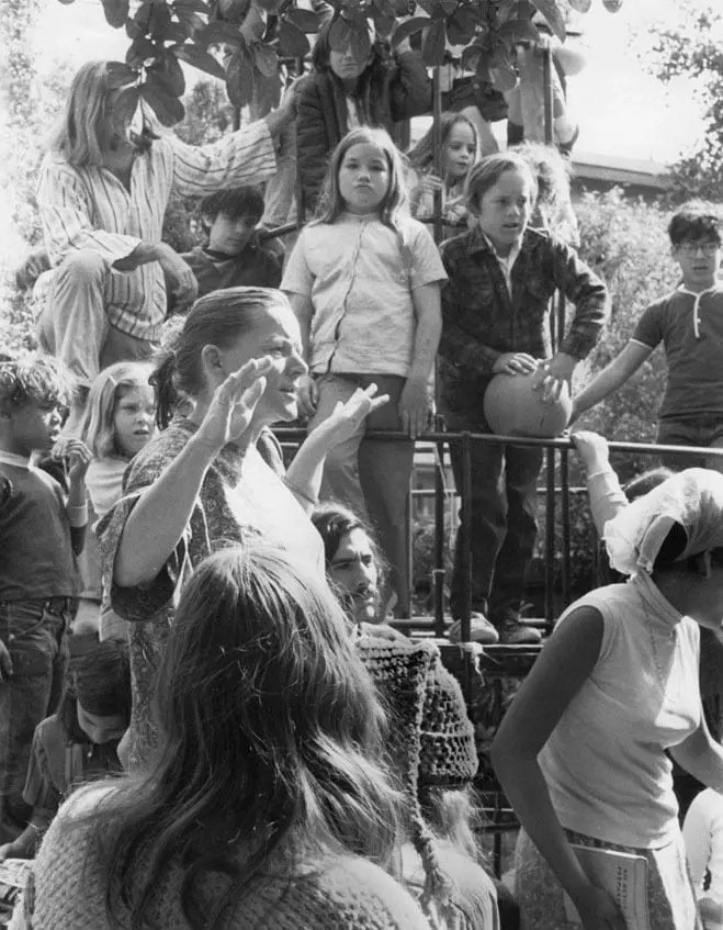 A woman gestures with raised hands while speaking to a group of children standing on and around a metal structure outdoors.