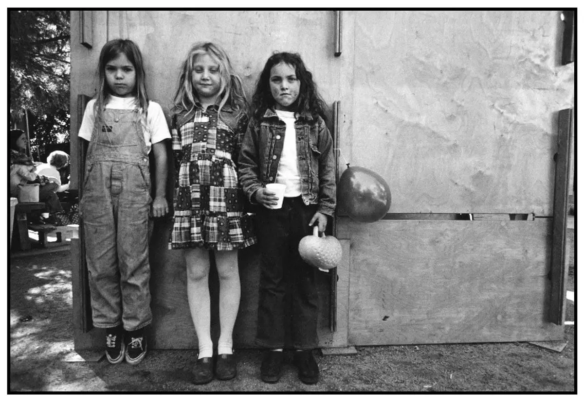 Three girls stand side by side against a plywood wall outdoors, one holding a paper cup and another holding a small decorated container, with a balloon tied nearby.