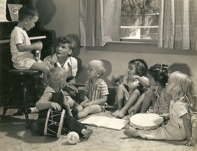 A black-and-white photo shows five children seated on the floor and one child sitting at a piano, with some holding small instruments.