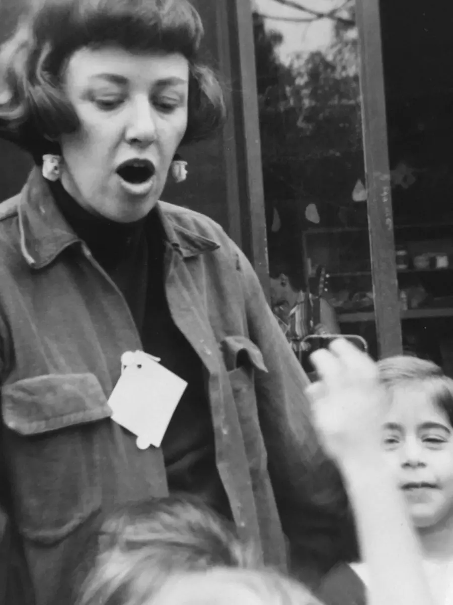 A woman with short hair and a name tag speaks with an open mouth while children look on, one raising a hand.