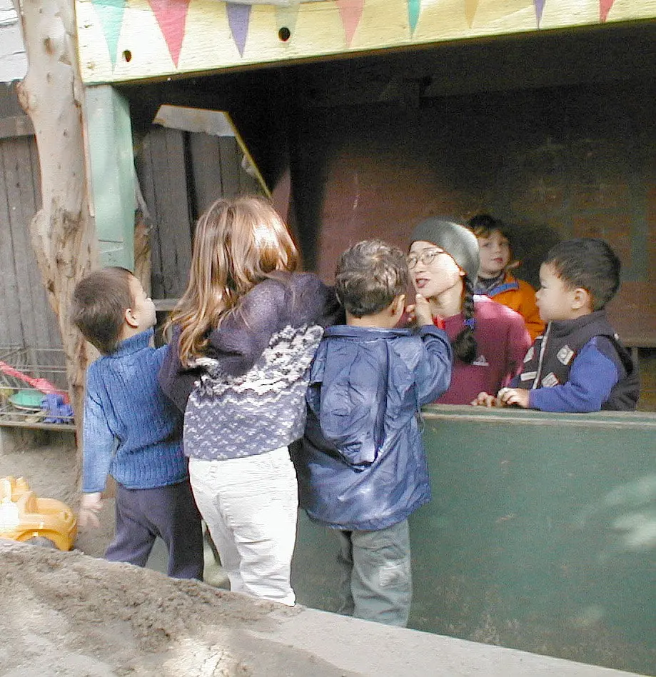 Children and an adult stand outside a wooden play structure with colorful bunting, with some children behind a green panel and others facing away in the foreground.