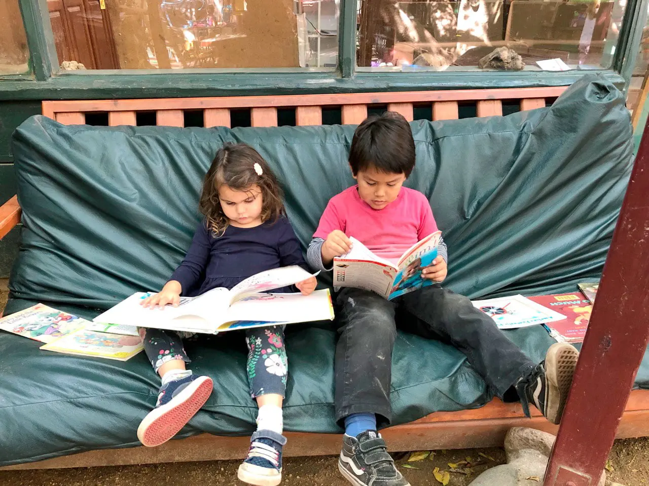 Two children sit on a green cushioned bench reading open books, with additional books resting on the seat beside them.