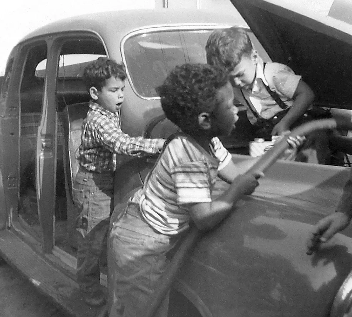 Three young boys climb into and around an open car, with one boy sitting inside the door, one leaning over the side, and one reaching into the open trunk.