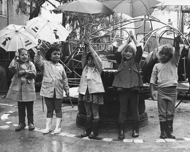 Five young children stand in a row on a wet playground surface, holding umbrellas or raising their arms, in front of a metal climbing structure.