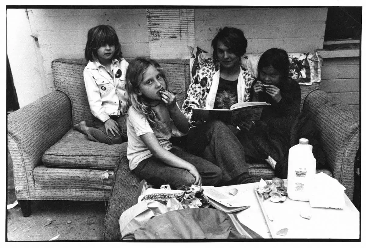 Four people sit on a woven couch, with one child eating, one holding an open book, and another eating near a table with a jug and food items.