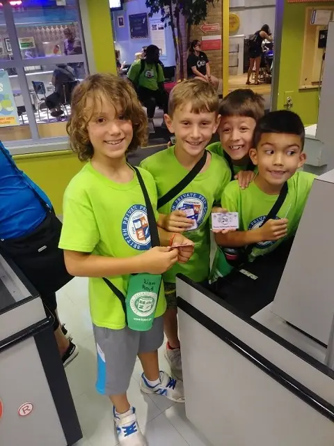 Four children in matching green shirts stand together holding small cards in an indoor play area with a play structure behind them.