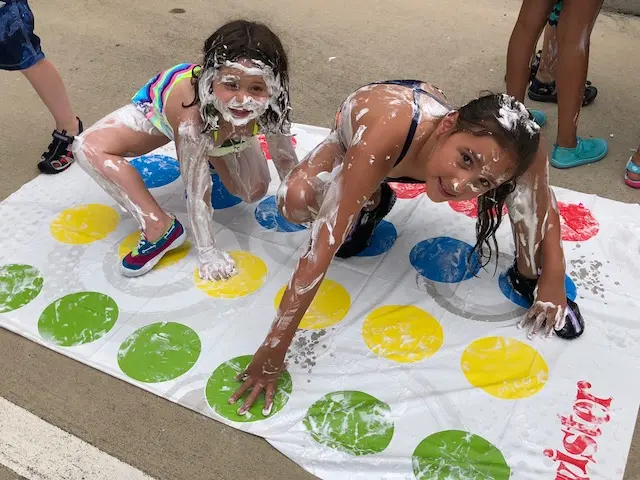 Two girls covered in white foam crawl on a white mat with colorful circles while other children stand nearby on pavement.