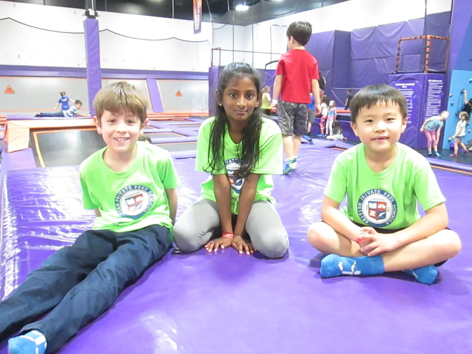 Three children wearing matching green shirts sit on a purple trampoline mat inside an indoor activity center while other children play in the background.