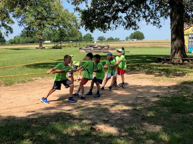 Five children in matching green shirts pull a yellow rope across a dirt patch in a grassy park area with trees and wooden play structures behind them.