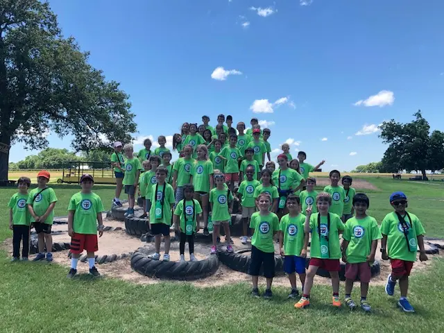 A large group of children in matching bright green t-shirts stands and sits on a pile of tires on a grassy field under a blue sky.