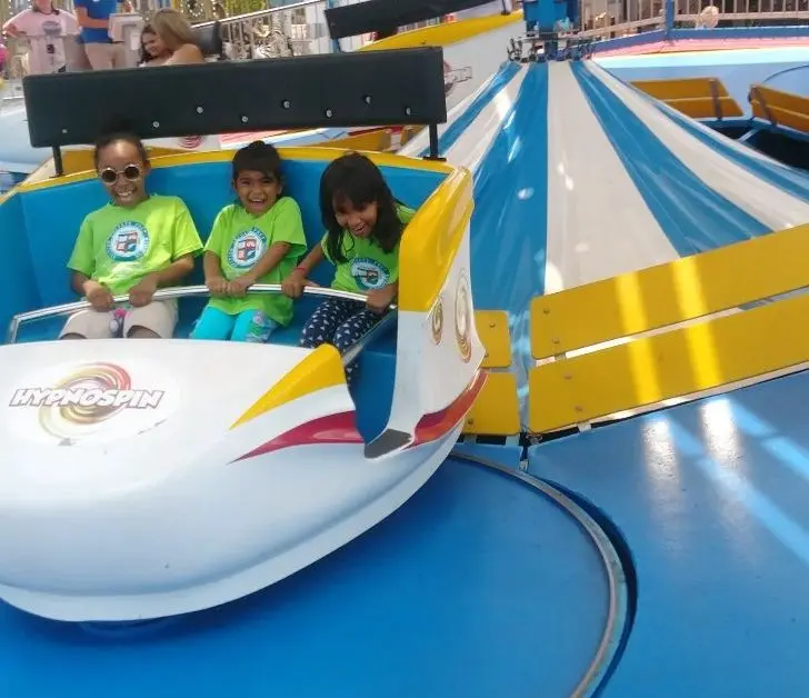 Three children sit in a blue and white amusement ride car labeled HypnoSpin, with two smiling at the camera and one looking down.