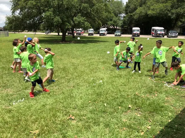 Children in matching green shirts throw white balls across a grassy field while others run and reach for them.