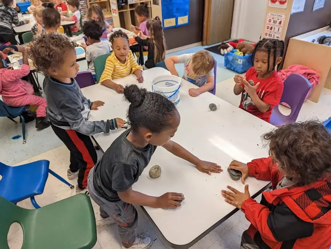 Several children stand and sit around a white table, handling small rocks and a bucket in a classroom setting.