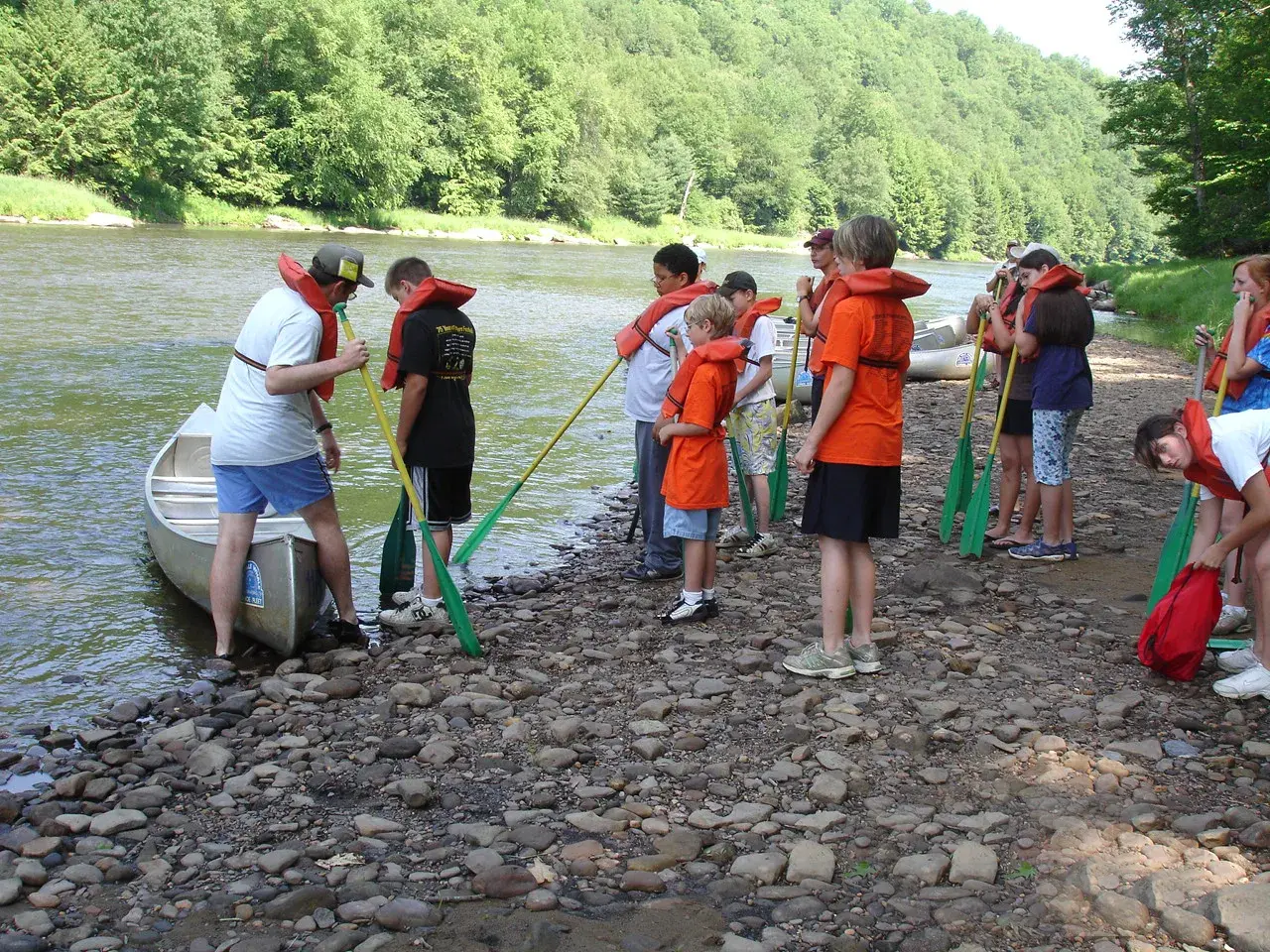 A group of children and adults wearing life jackets stand on a rocky riverbank holding paddles near a canoe and kayaks.