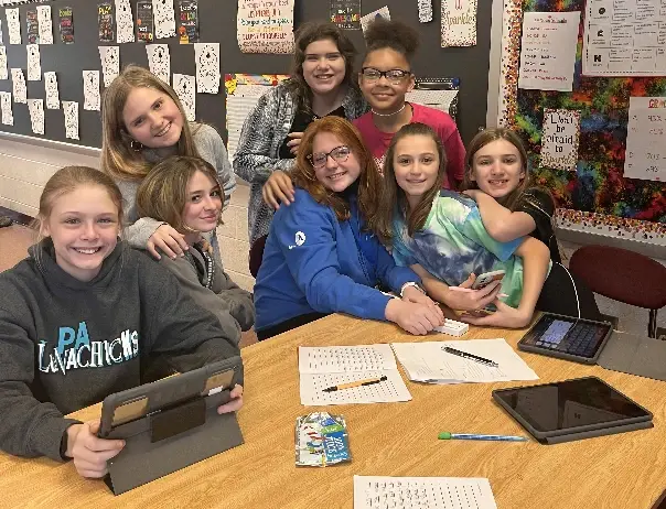 Eight girls sit and stand around a wooden table with tablets, papers, and a snack wrapper in a classroom with a bulletin board behind them.