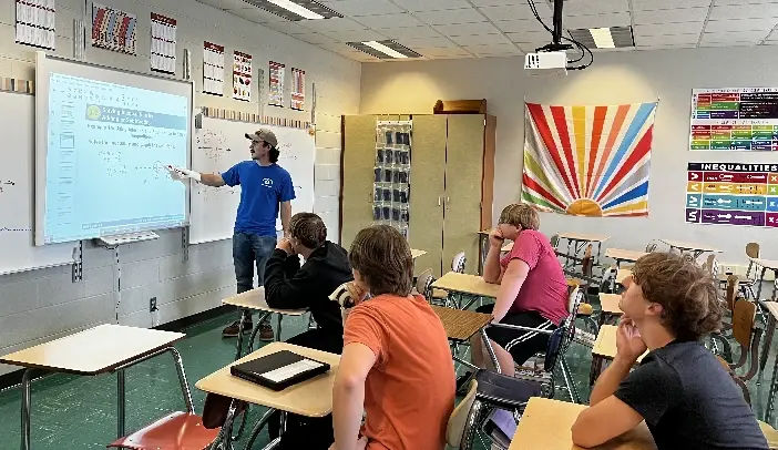 A teacher in a blue shirt points at a projected screen while four students sit at desks in a classroom with colorful wall displays.