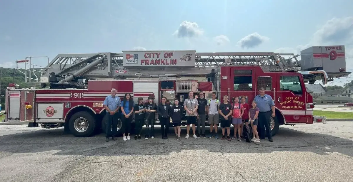 A group of children and two adults stand in a line in front of a red fire ladder truck parked on pavement under a cloudy sky.