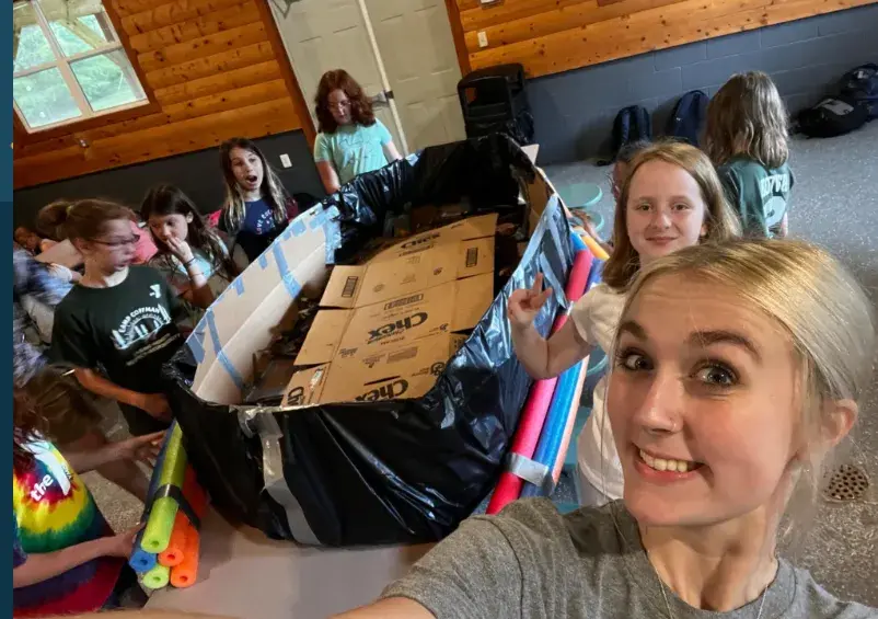 A girl in the foreground takes a selfie with a group of children gathered around a large cardboard box lined with a black plastic bag and pool noodles in a wood-paneled room.