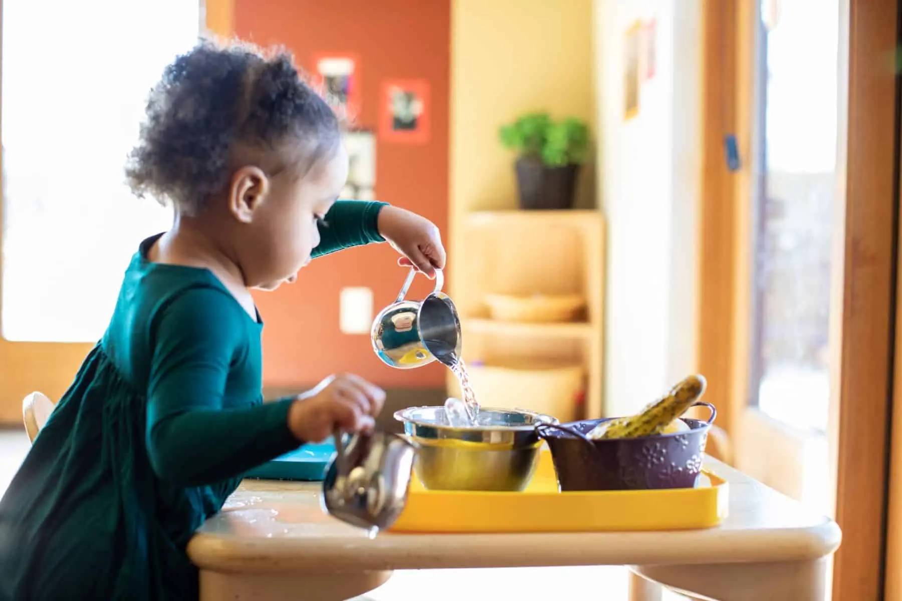 A young child in a green dress pours water from a small metal pitcher into a bowl on a yellow tray holding bowls and a cucumber.