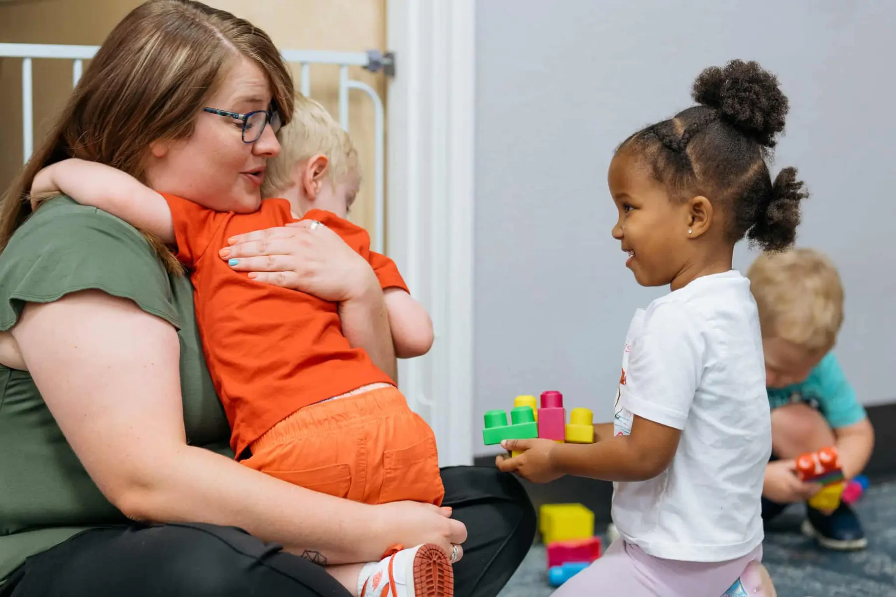 A woman sits on the floor hugging a child in an orange shirt while a girl holding colorful blocks stands nearby and another child plays with blocks in the background.