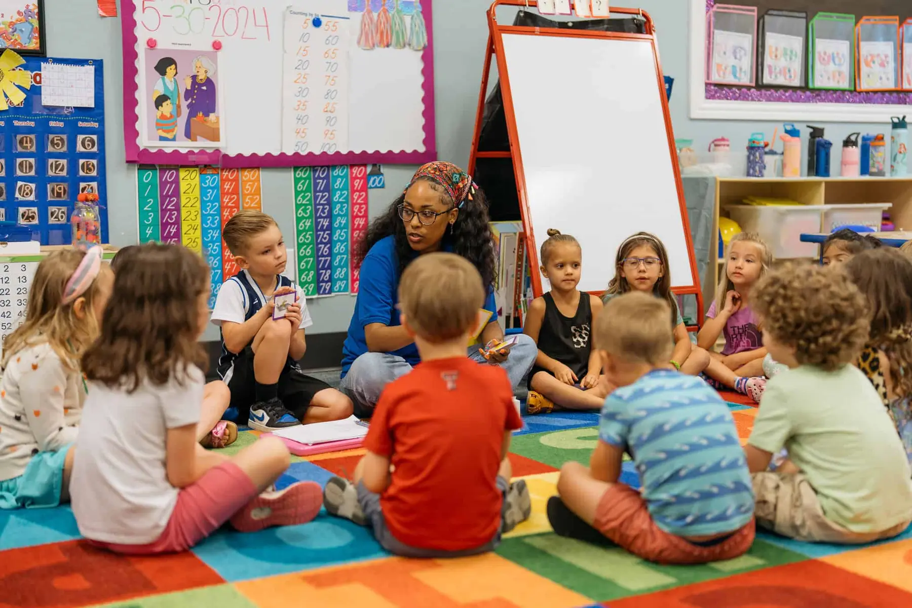 A woman sits on a colorful rug speaking to a group of children seated around her in a classroom with number charts and a whiteboard.