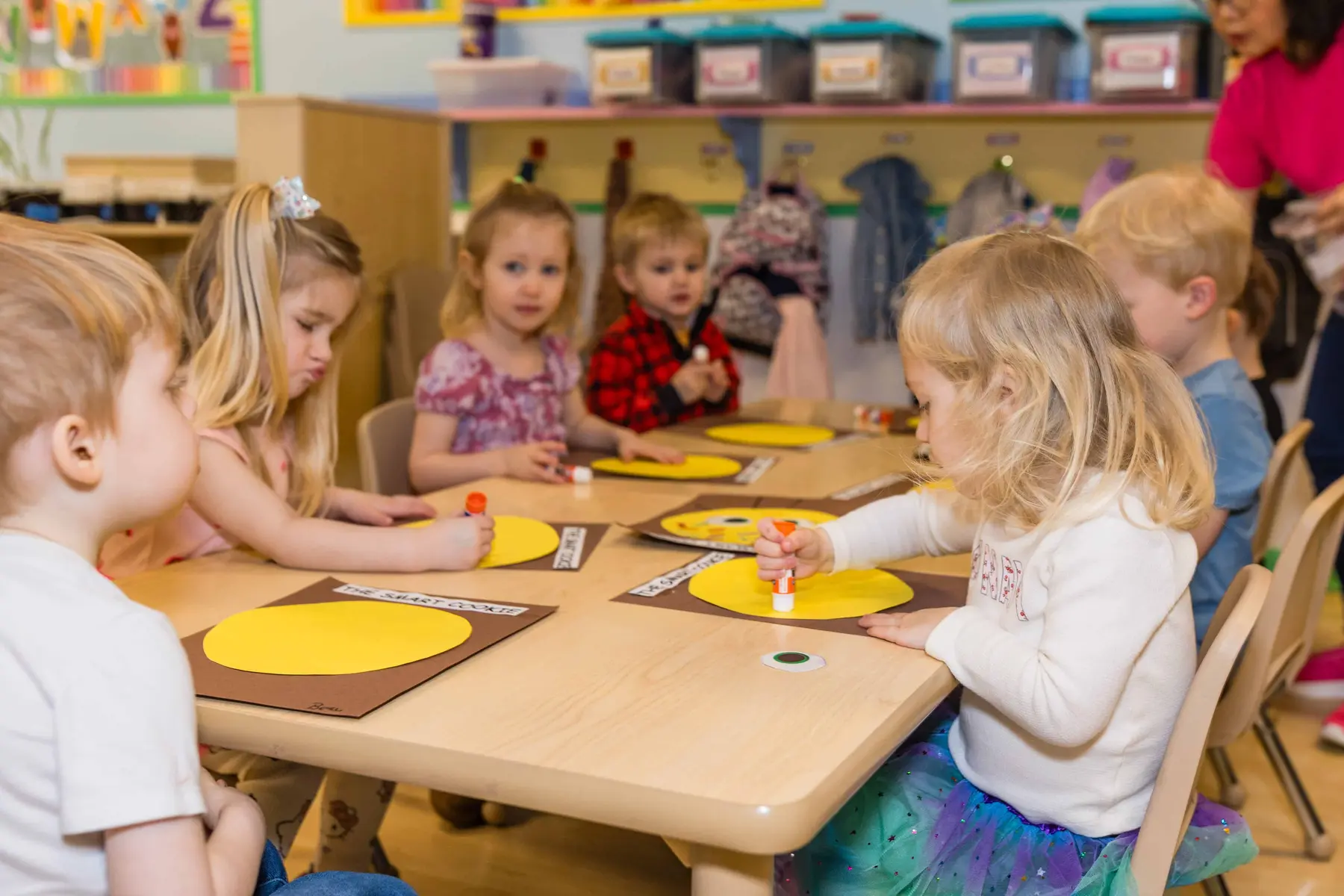 Several young children sit at a table painting yellow paper circles on brown mats while an adult stands in the background.
