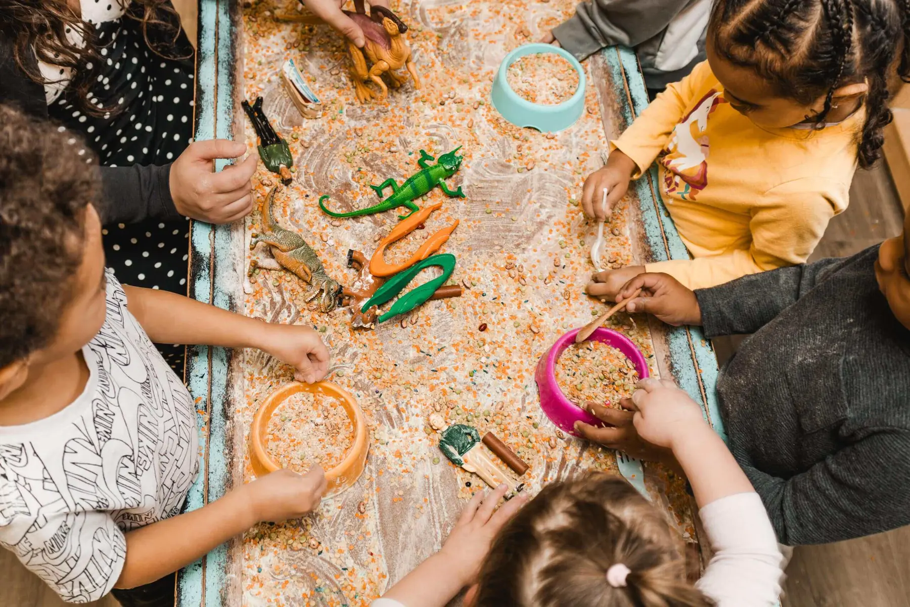 Five children stand around a shallow tray filled with seeds and toy reptiles, using bowls and scoops to play.