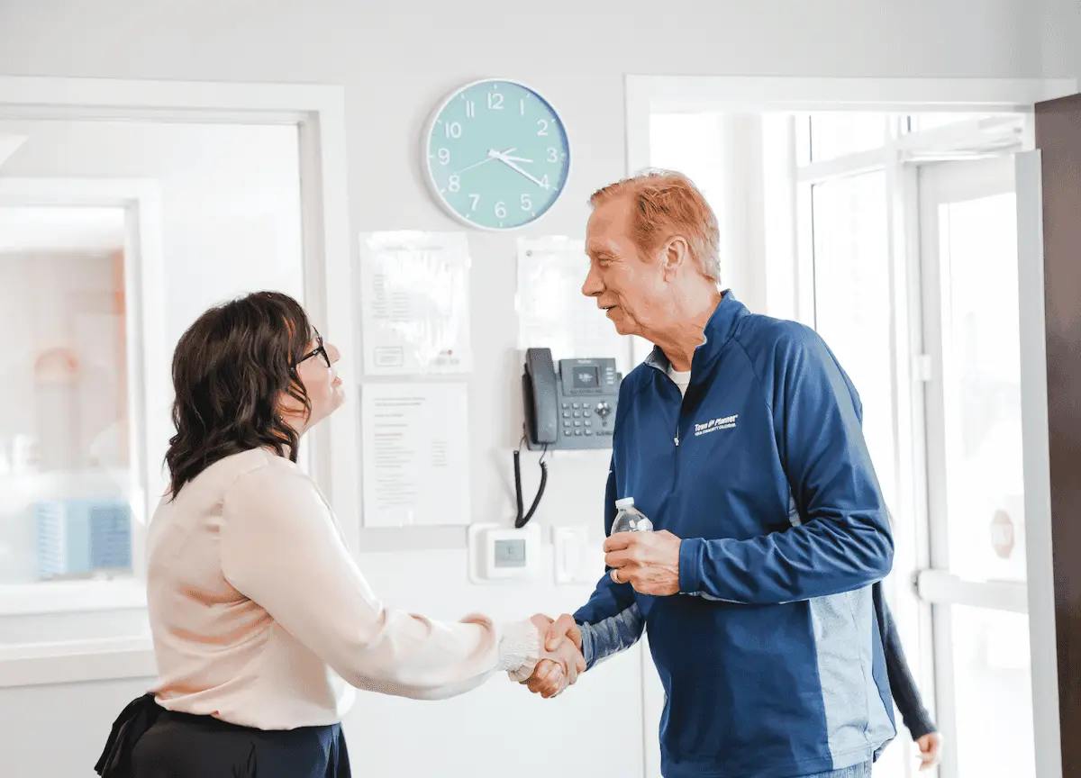 A woman in a light blouse and dark skirt shakes hands with an older man in a blue jacket holding a water bottle, standing near a wall with a clock, papers, and a phone.