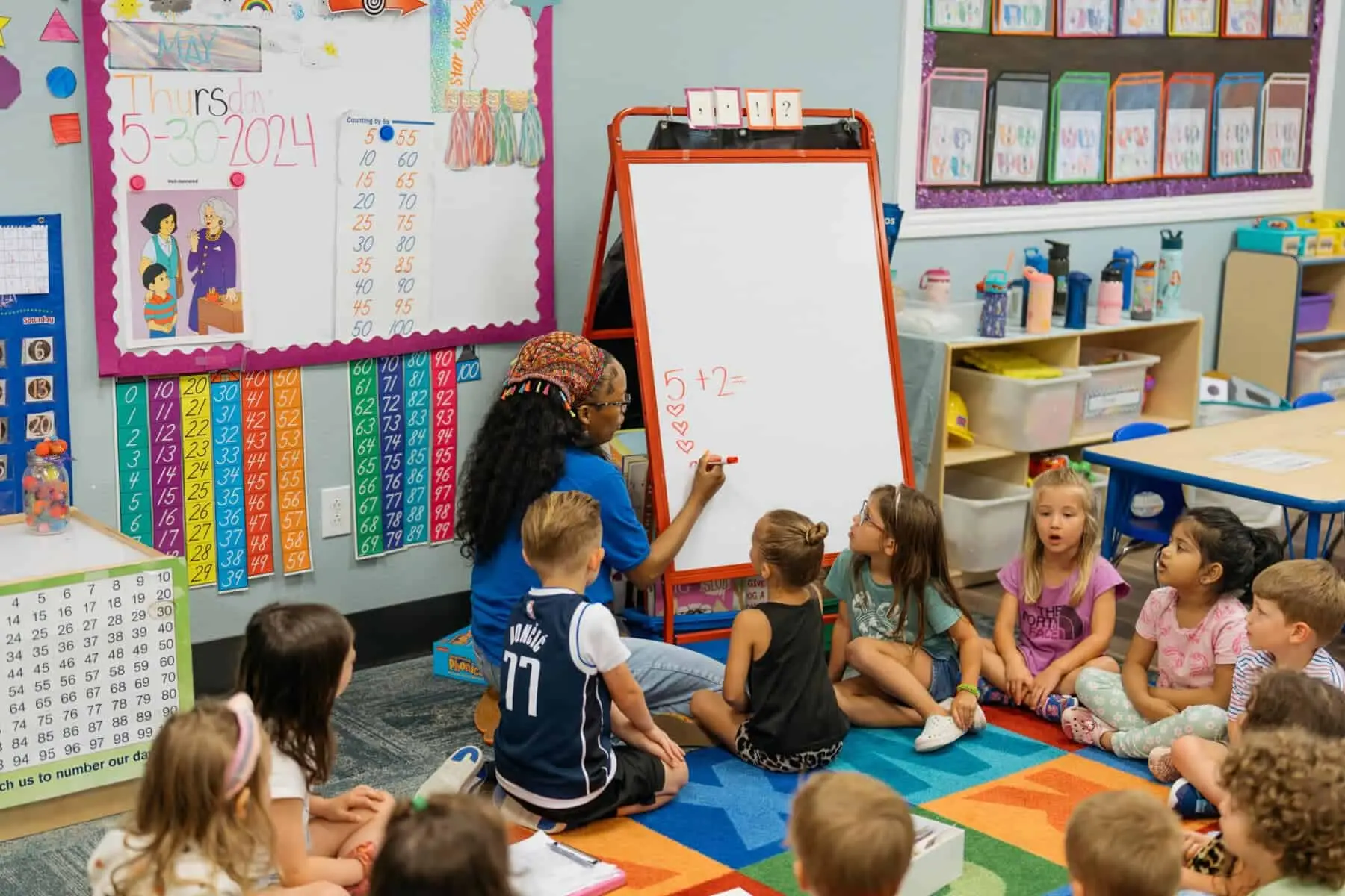 A teacher writes a math problem on a whiteboard while a group of children sit on a colorful rug and watch.