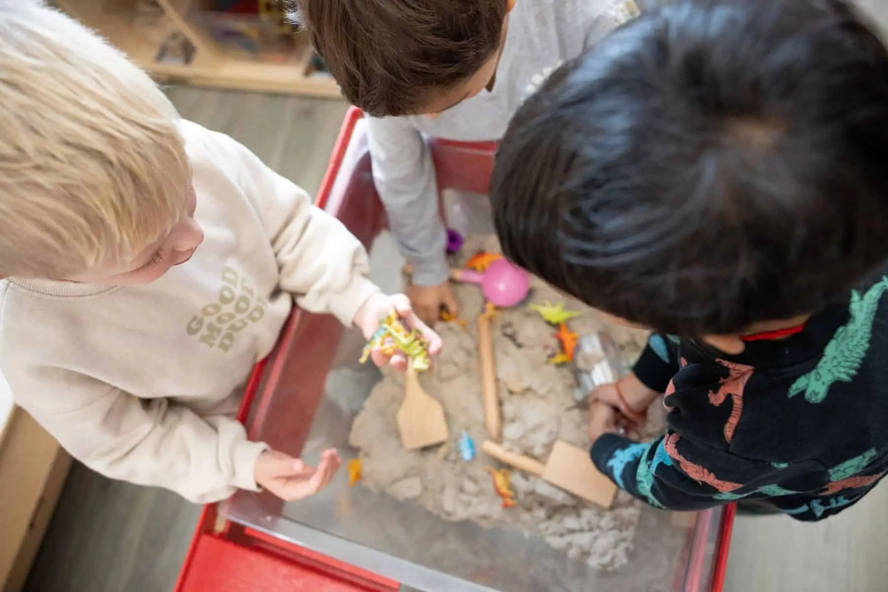 Three children stand around a red sand table, playing with toy dinosaurs and wooden scoops in the sand.