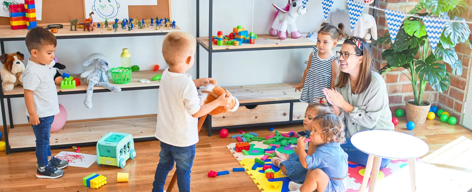 Children and an adult play with toys on a rug in a room with shelves of toys and a plant.