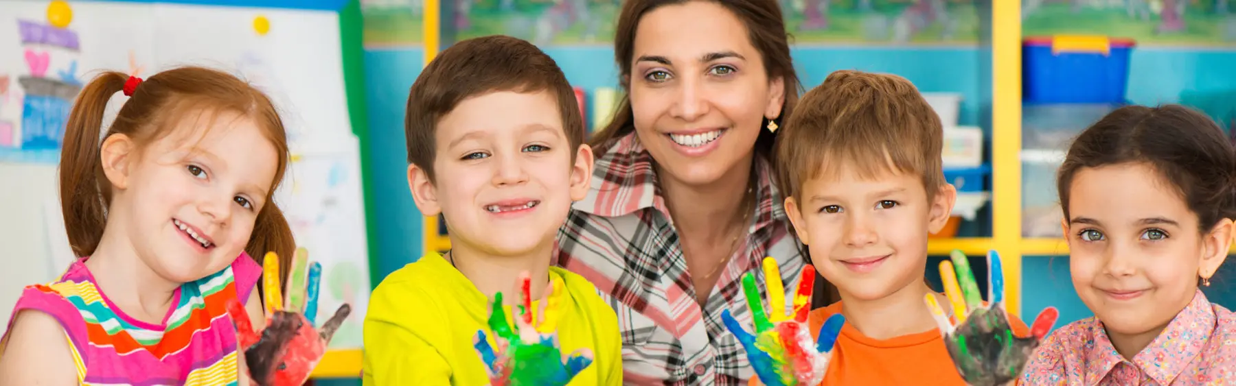 Four children and an adult woman smile at the camera while showing painted hands, with the children wearing colorful tops and the woman in a plaid shirt.