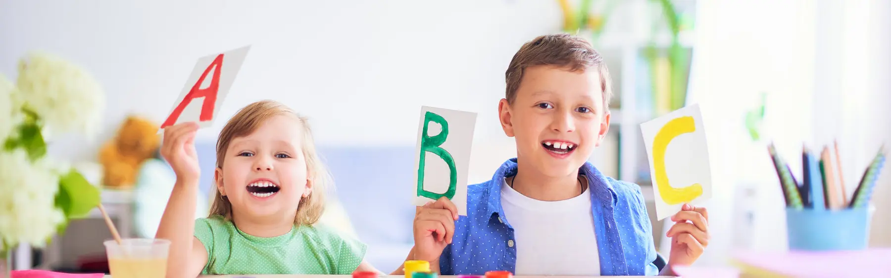 A boy and a girl sit at a table holding up letter cards A, B, and C while smiling at the camera.