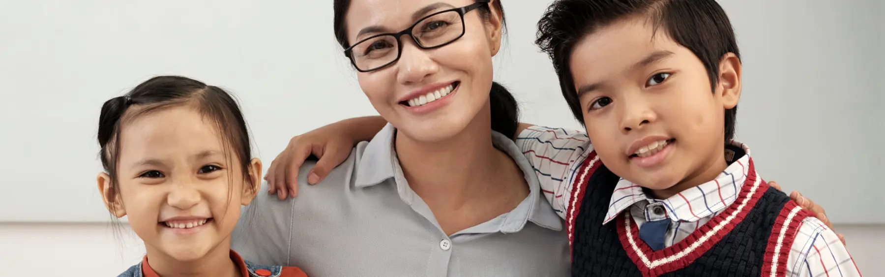 A woman with glasses smiles between a young girl and a boy in a sweater vest, all facing the camera.