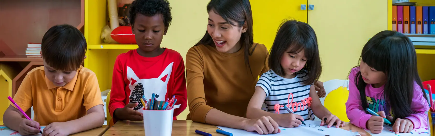 Four children and an adult sit at a table drawing and coloring with markers and pencils in a yellow-walled room.