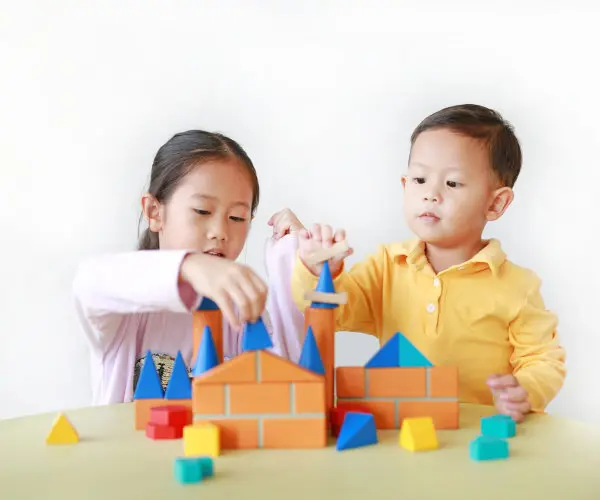 Two children sit at a table and stack colorful foam building blocks into a castle shape.