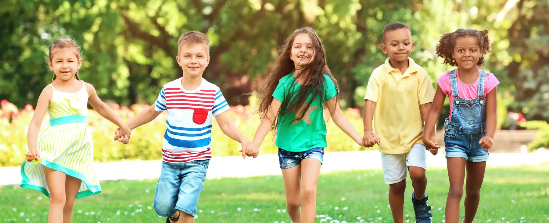 Five children hold hands in a line while walking on grass in a park with trees behind them.