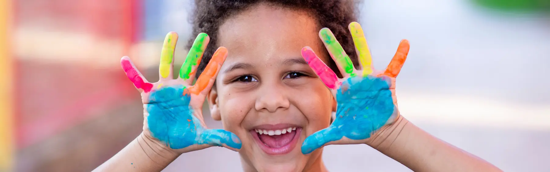 A smiling child holds up two hands covered in bright blue, green, orange, and pink paint.