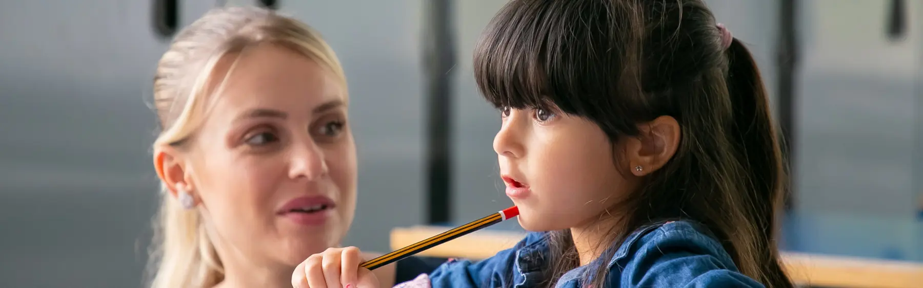 A woman looks toward a girl who holds a pencil near her mouth while wearing a blue denim top.